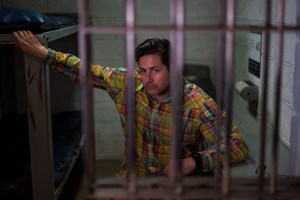 A white man with thick dark hair in a checkered colourful shirt poses behind the bars of a prison cell